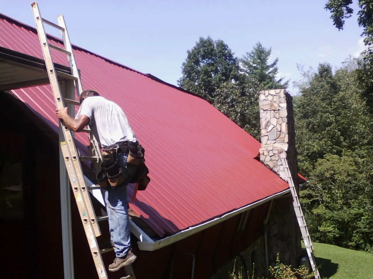 Roofer on a ladder beside a red metal roof and stone chimney — Soffit & Fascia Repair by Roofing Brothers in Groves